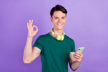 Sticker - Photo of brunette positive young man hold phone okay sign headphones isolated on violet color background