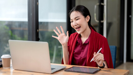 Wall Mural - Beautiful young Asian businesswoman wearing a red dress at work via video call using laptop holding pen tablet at office.