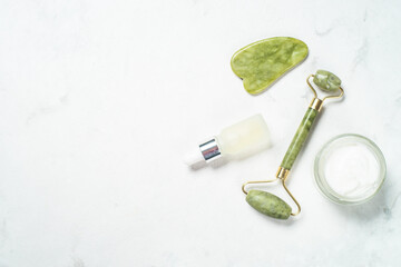 Fototapete - Face massage set on white background. Jade roller and gua sha massagers with face cream and serum bottle. Top view.