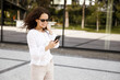 © Dragosh - Smiling young woman, close-up portrait, looking at the cell phone, against a building with window background.
