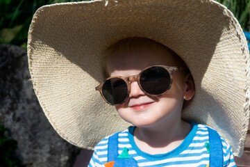 Cute little toddler wearing an oversized sun hat and sunglasses relaxing in the sun