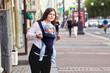 © grigvovan - Young woman with baby in sling stands on crowded street in city.