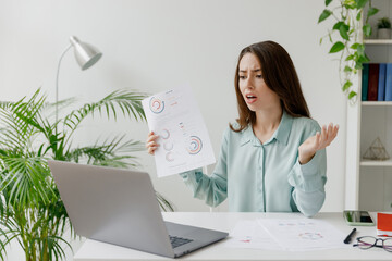 Young sad confused employee business woman in blue shirt hold paper account documents sit work at workplace white desk advises online on laptop pc computer at office indoor Achievement career concept