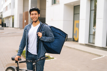 Wall Mural - Medium shot portrait of joyful handsome young delivery man in protective helmet posing standing near bicycle in city street, looking at camera. Courier male with backpack delivery food to client