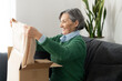 © Vadim Pastuh - Happy senior mature adult woman is looking at her new beige jumper, sitting on the couch, and unpacking parcel. Smiling online lady opening online store order in the postal delivery shipping box