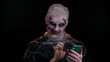 Frightening man with Halloween zombie bloody wounded makeup using mobile phone typing new post on web, sms message, browsing, addiction of social networks. Sinister undead guy on black wall background