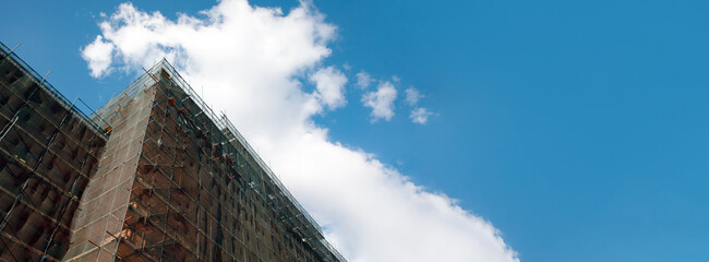 Building facade renovation over blue sky, old house reconstruction, repair. Scaffold in front of building facade covered with transparent fabric, panoramic layout
