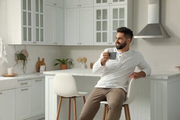 Wall Mural - Handsome young man with cup of tea sitting on stool in kitchen