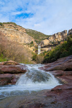 Small Waterfall Between Red Rocks With Cliff In The Background With Monastery, Sant Miquel Del Fai Catalonia, Spain