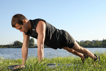 Canvas Print - Sporty man doing straight arm plank exercise on green grass near river