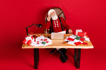 Full size photo of santa claus happy positive smile dream think sit table workshop noel isolated over red color background