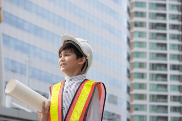 Little boy wearing a engineering helmet and holding blueprint paper for construction in the city. Inspiring future for children concept.