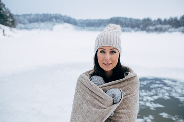 Winter swimming. Woman ready to swim in ice water. How to swim in cold water. Beautiful young woman wrapped in a towel and swimming clothes. Gray hat and gloves. People and nature lake in the forest.