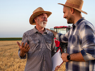 Poster - Two male farmers standing in harvested field talking in front of tractor.