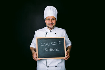 Cook dressed in a white hat and jacket holds a blackboard that reads: Cooking School on a black background. Restaurant, food and cooking school concept.