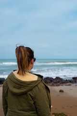 Sticker - A woman in a winter coat admiring the view of the ocean with waves crashing on a sandy beach