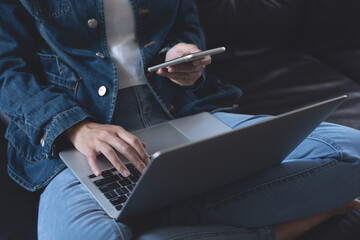 Wall Mural - Young casual woman, freelancer in blue jeans and sweater sitting on sofa using mobile phone, working on laptop computer at home, close up