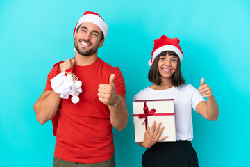 Wall Mural - Young couple with christmas hat handing out gifts isolated on blue background giving a thumbs up gesture because something good has happened