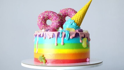 Wall Mural - Cake on birthday with multicolored rainbow cream on a yellow background decorated with a waffle cone, pink donuts, sprinkles, marmalade candies poured with chocolate.