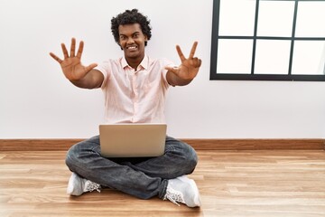 Poster - African man with curly hair using laptop sitting on the floor showing and pointing up with fingers number eight while smiling confident and happy.