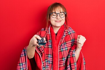 Poster - Redhead young woman wrapped in a red warm red blanket using tv control screaming proud, celebrating victory and success very excited with raised arm