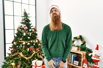 Wall Mural - Redhead man with long beard wearing christmas hat by christmas tree looking at the camera blowing a kiss on air being lovely and sexy. love expression.