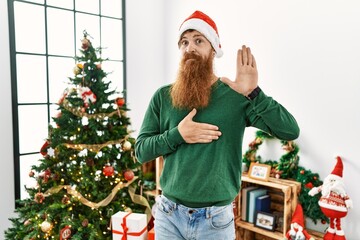 Poster - Redhead man with long beard wearing christmas hat by christmas tree swearing with hand on chest and open palm, making a loyalty promise oath