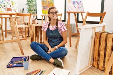 Wall Mural - Young brunette woman at art studio sitting on the floor looking away to side with smile on face, natural expression. laughing confident.