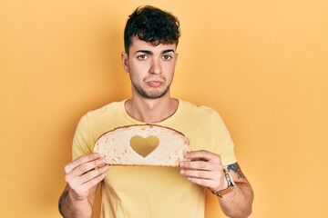 Sticker - Young hispanic man holding bread with heart shape over eye depressed and worry for distress, crying angry and afraid. sad expression.