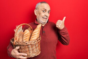 Poster - Handsome mature man holding wicker basket with bread pointing thumb up to the side smiling happy with open mouth