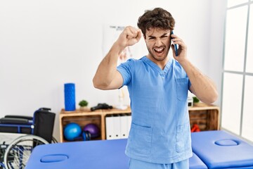 Sticker - Hispanic physiotherapy man working at pain recovery clinic speaking on the phone annoyed and frustrated shouting with anger, yelling crazy with anger and hand raised