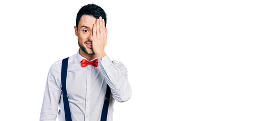 Poster - Hispanic man with beard wearing hipster look with bow tie and suspenders covering one eye with hand, confident smile on face and surprise emotion.