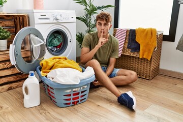 Sticker - Young caucasian man putting dirty laundry into washing machine asking to be quiet with finger on lips. silence and secret concept.