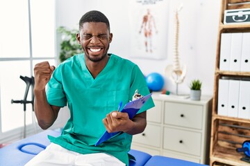 Sticker - Young african american man working at pain recovery clinic very happy and excited doing winner gesture with arms raised, smiling and screaming for success. celebration concept.