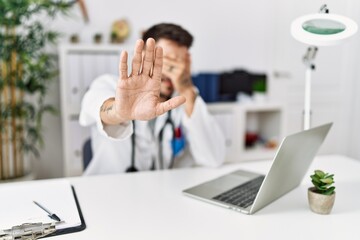 Poster - Young doctor working at the clinic using computer laptop covering eyes with hands and doing stop gesture with sad and fear expression. embarrassed and negative concept.