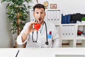 Poster - Young doctor holding heart at medical clinic scared and amazed with open mouth for surprise, disbelief face