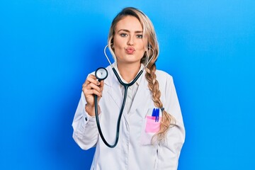 Wall Mural - Beautiful young blonde doctor woman holding stethoscope looking at the camera blowing a kiss on air being lovely and sexy. love expression.