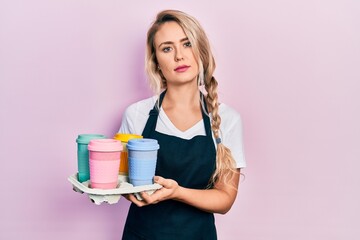 Poster - Beautiful young blonde woman wearing waitress apron holding take away cup of coffees relaxed with serious expression on face. simple and natural looking at the camera.