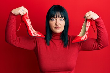 Poster - Young hispanic woman holding elegant red heels relaxed with serious expression on face. simple and natural looking at the camera.