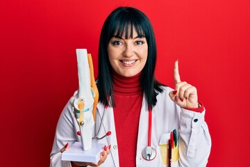 Sticker - Young hispanic doctor woman holding anatomical model of knee joint smiling with an idea or question pointing finger with happy face, number one