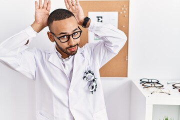 Canvas Print - African american optician man standing by eyesight test doing bunny ears gesture with hands palms looking cynical and skeptical. easter rabbit concept.