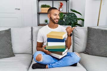 Poster - African american young man holding a pile of books sitting on the sofa smiling with happy face looking and pointing to the side with thumb up.