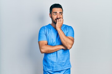 Canvas Print - Handsome hispanic man with beard wearing blue male nurse uniform looking stressed and nervous with hands on mouth biting nails. anxiety problem.