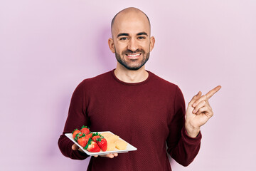 Sticker - Young bald man eating fresh and healthy fruits smiling happy pointing with hand and finger to the side