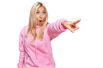 Wall Mural - Young blonde woman wearing casual sweatshirt pointing with finger surprised ahead, open mouth amazed expression, something on the front