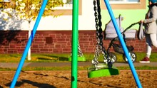 A Green Swing With Chains Swings Back And Forth And A Woman Rolls A Carriage With A Newborn Baby Behind It