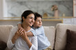 © fizkes - Dreamy bonding sincere small mixed race kid boy cuddling indian young mother, looking in distance, visualizing future or remembering good moments, resting on cozy sofa together at home.