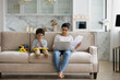 © fizkes - Focused young indian woman working distantly on computer or managing household budget while little kid son playing drawing on toy tablet, sitting together on cozy sofa, parenting and career concept.