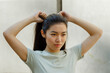 Pretty Asian young woman in t-shirt adjusts ponytail made of long dark hair standing near white wall on sunny day close view