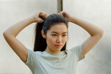 Fototapeta  - Pretty Asian young woman in t-shirt adjusts ponytail made of long dark hair standing near white wall on sunny day close view